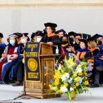 photo of Timnit Gebru in graduation regalia standing at a podium smiling with I School faculty seated behind her